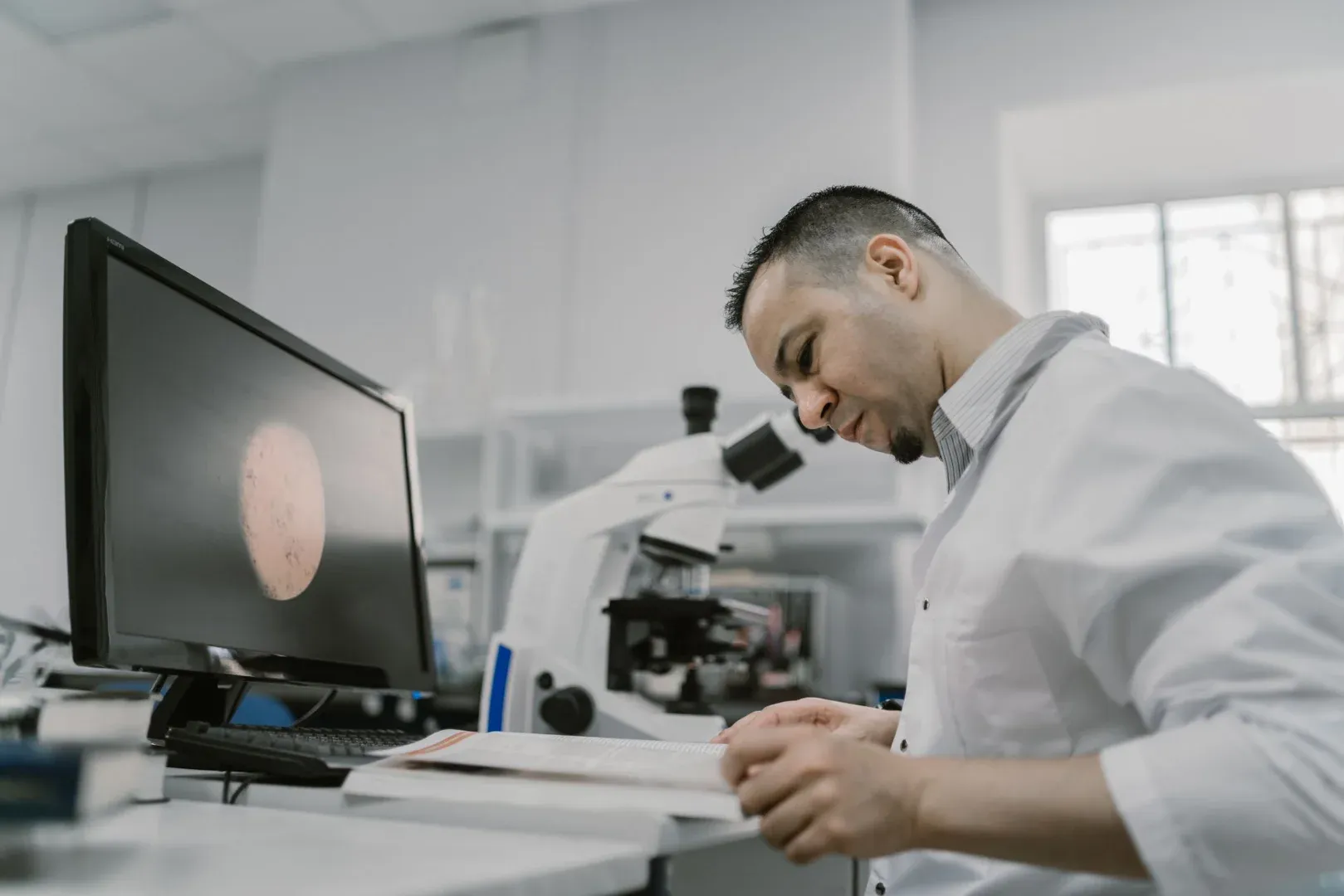 Asian man reading in a laboratory, surrounded by equipment and microscope, focused on work.