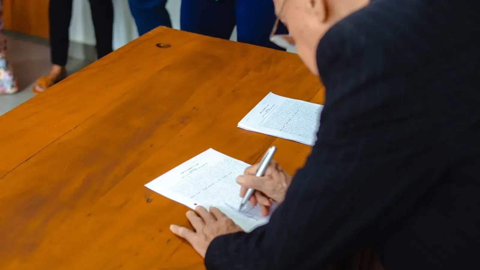 A close-up of a businessman signing official documents at a wooden desk.