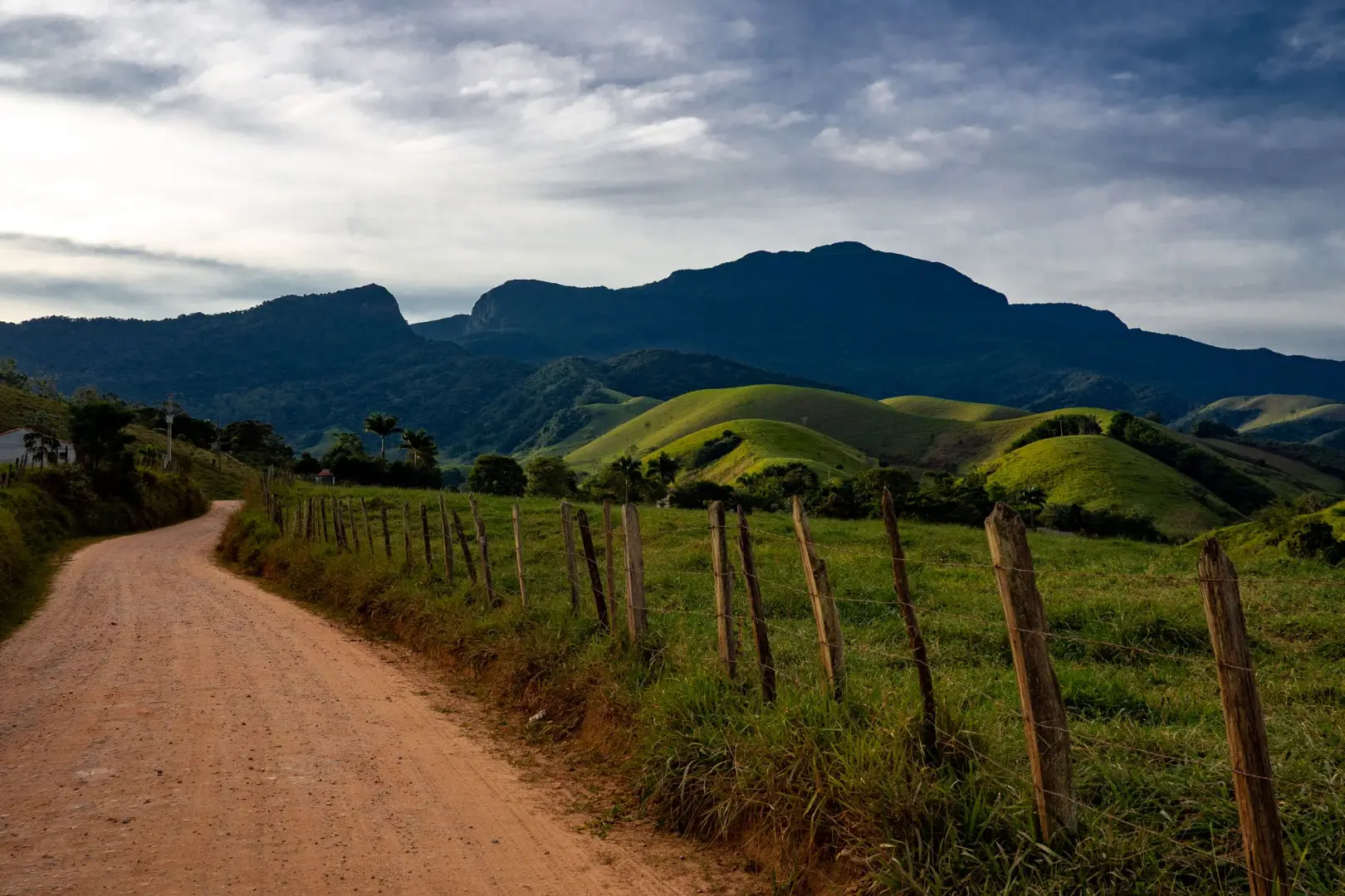 Captura de un camino de tierra a través de verdes colinas que conducen a una cadena montañosa bajo un cielo nublado.