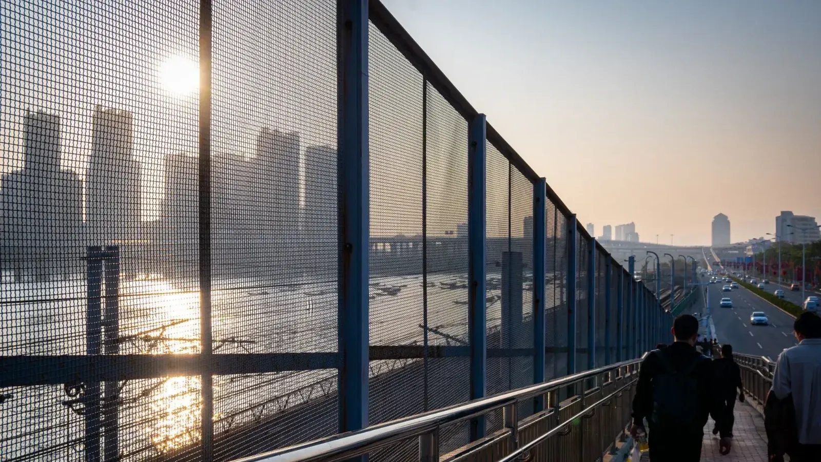 Silhouetted cityscape during sunset with people walking along a fenced bridge.