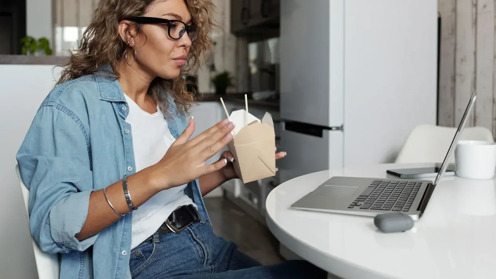 Una mujer enfocada en un ambiente acogedor en el hogar, trabajando en una computadora portátil mientras disfruta de la comida para llevar.