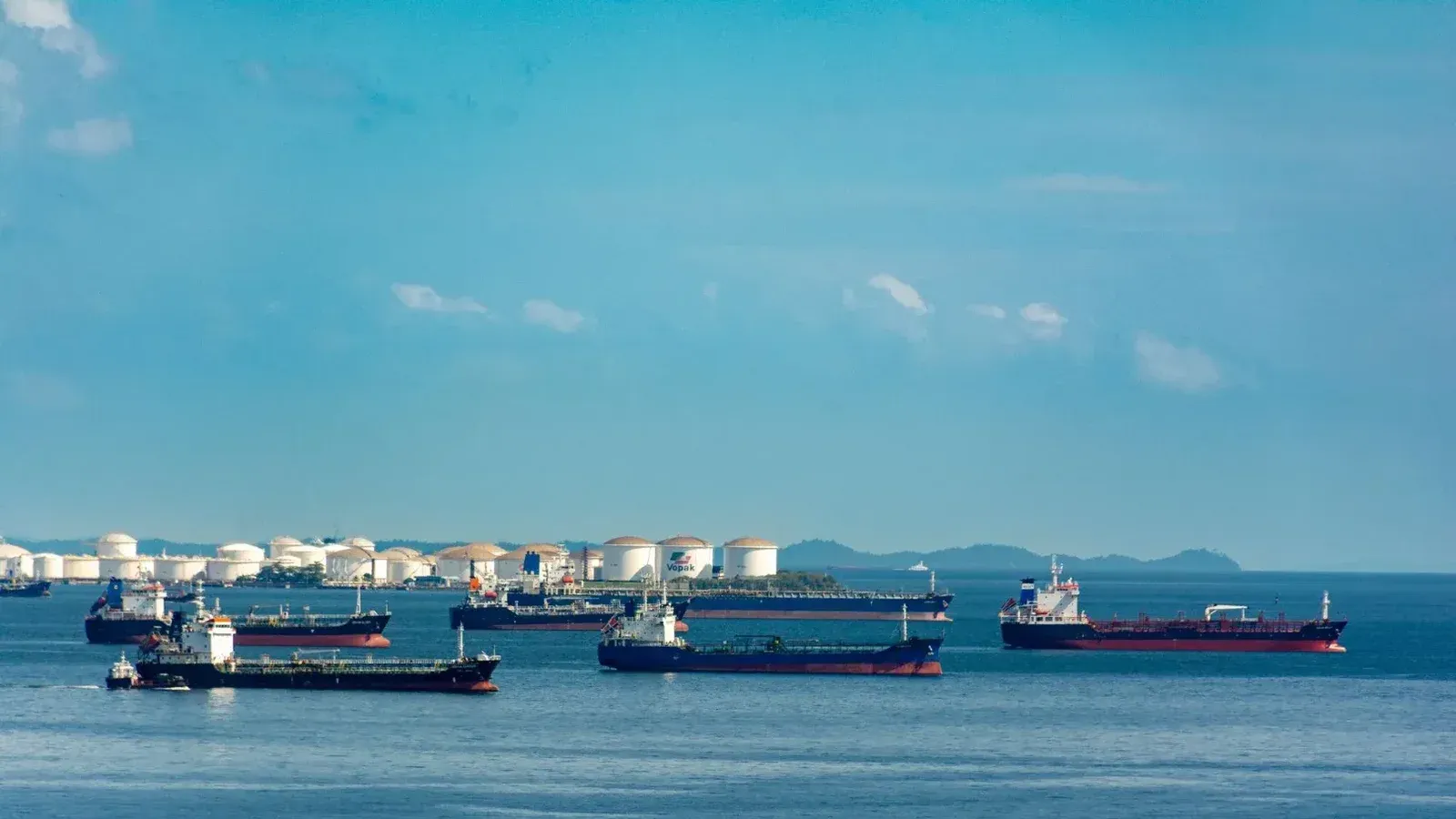 A fleet of cargo ships docked near oil storage tanks along a serene coastline with a clear blue sky above.