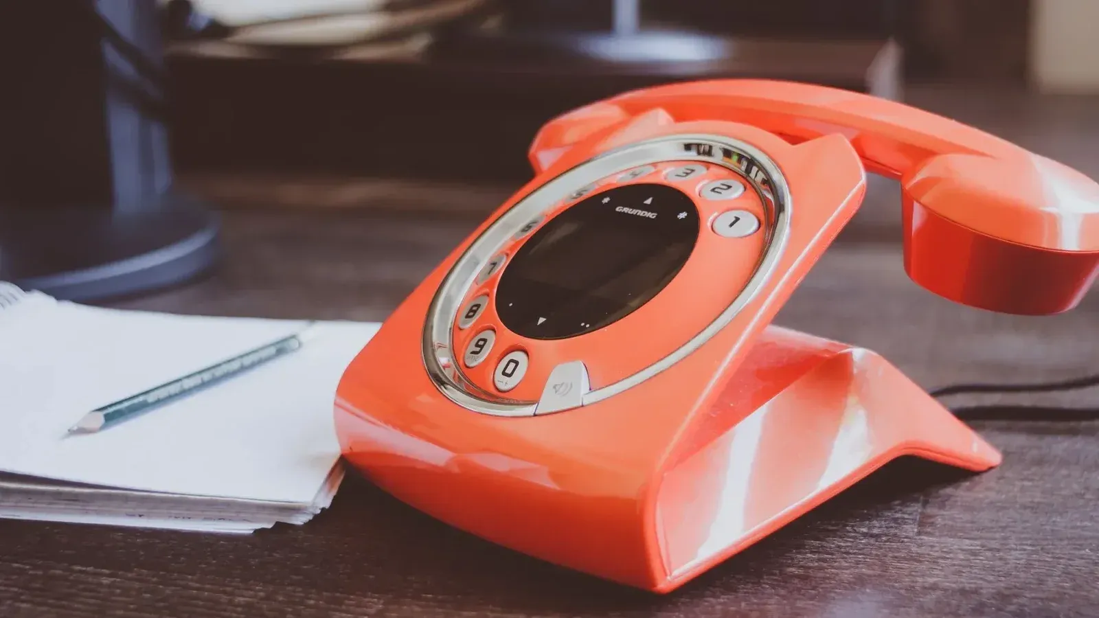 Un teléfono giratorio naranja de estilo retro se encuentra en un escritorio de madera junto con un lápiz y un bloc de notas.