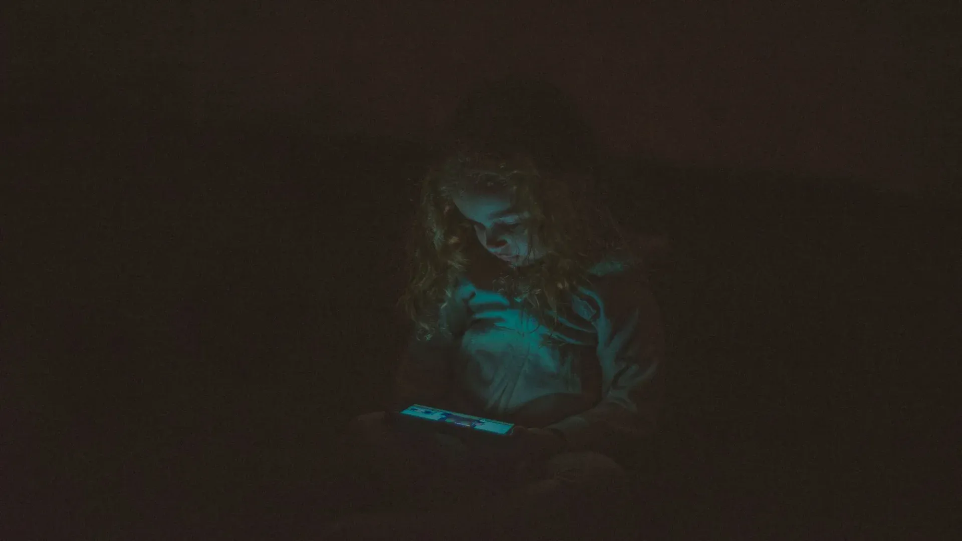 Focused little girl with long curly hair in casual clothes sitting and using smartphone while watching video in dark room