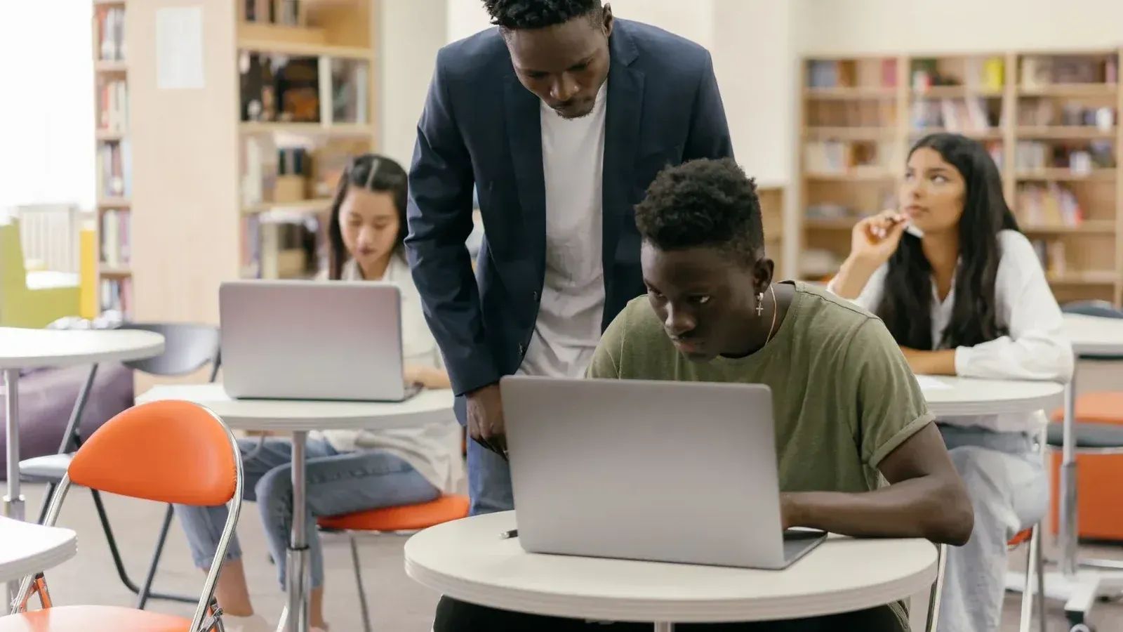 Students learning in a classroom setting with a teacher assisting and laptops on desks, creating an interactive education environment.