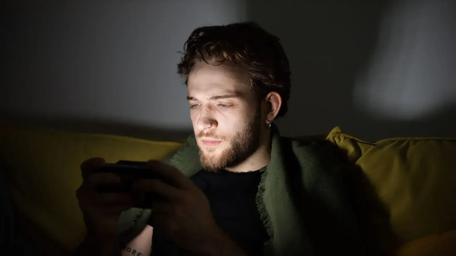 Un hombre barbudo jugando videojuegos en el interior, enfocado en una consola de mano.