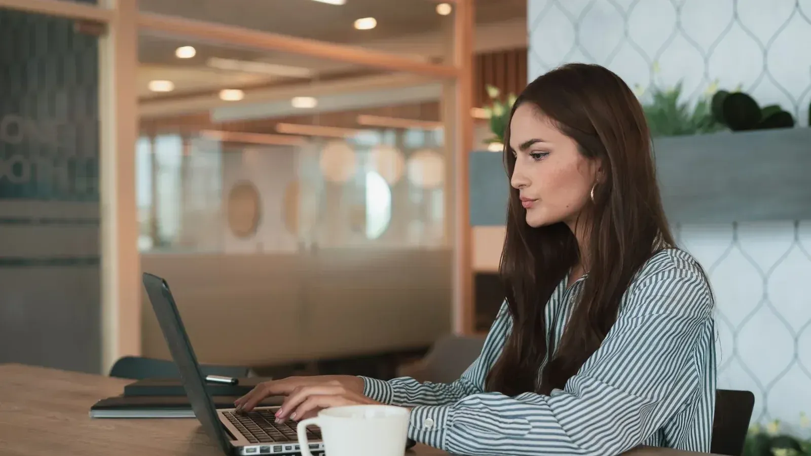A professional woman working on her laptop at a modern office with a coffee cup nearby.