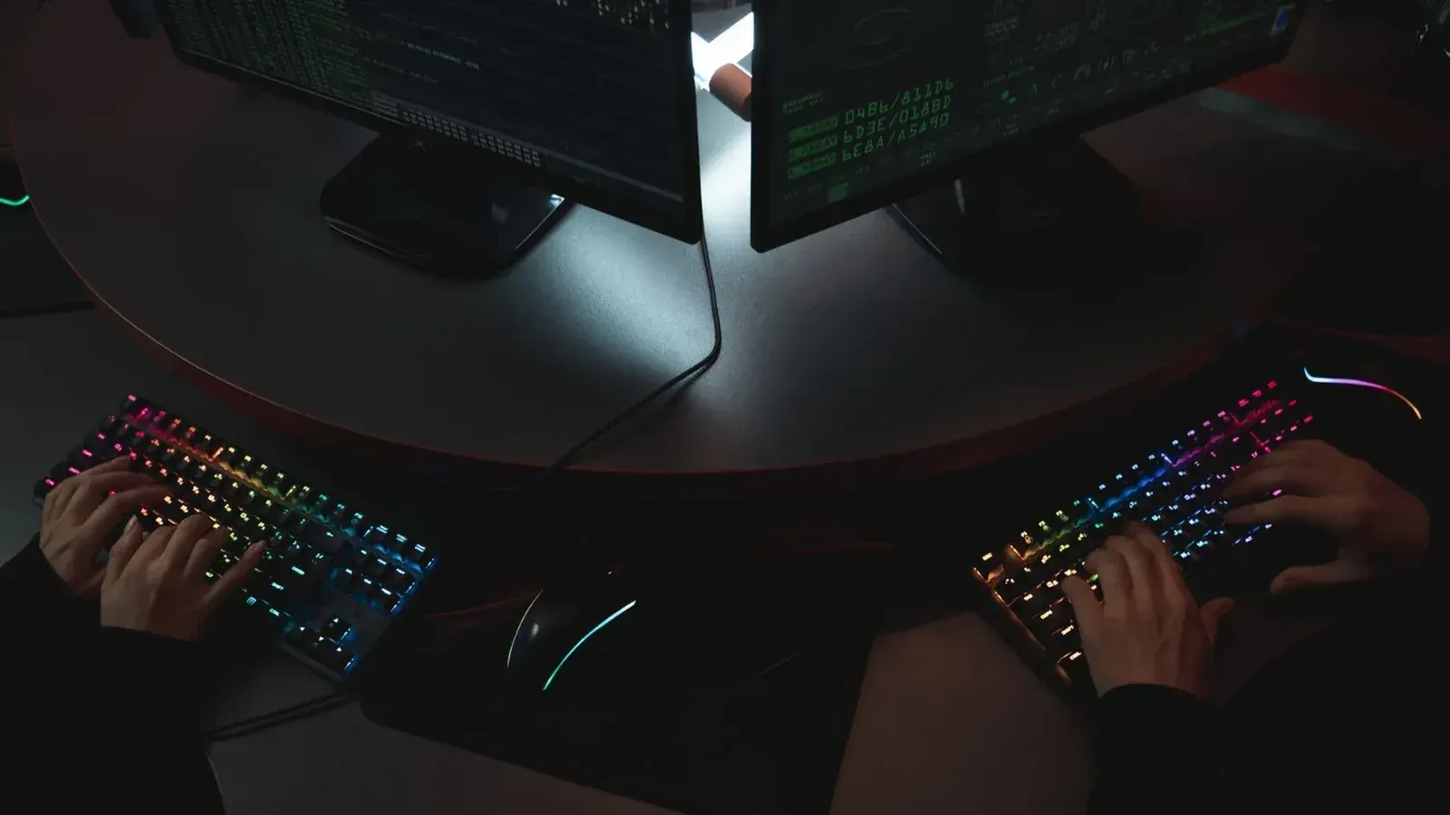Two people typing on RGB keyboards with code on screens, indicating a cybersecurity environment.