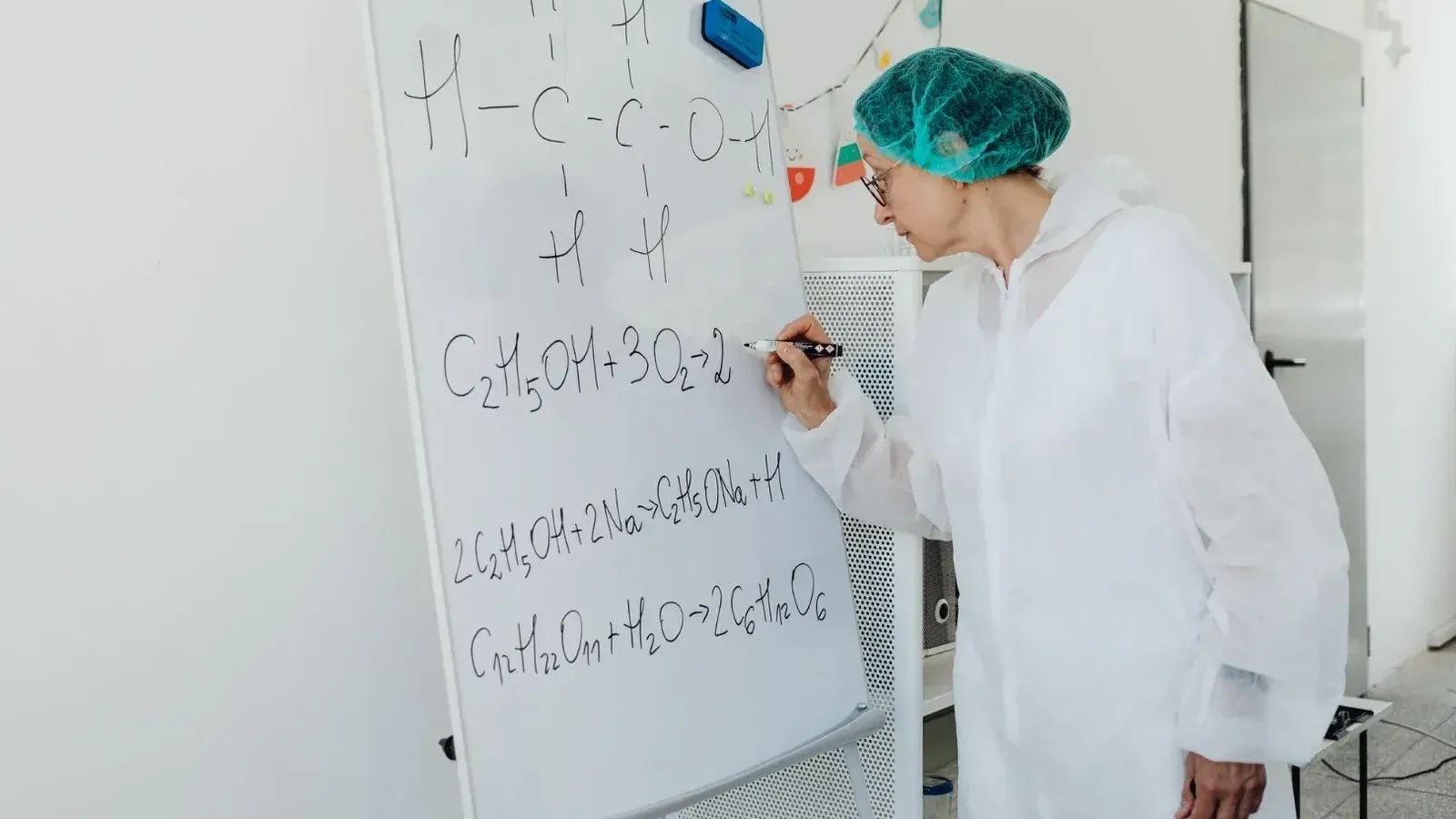 Anciana en bata de laboratorio escribiendo ecuaciones químicas en una pizarra en el interior.