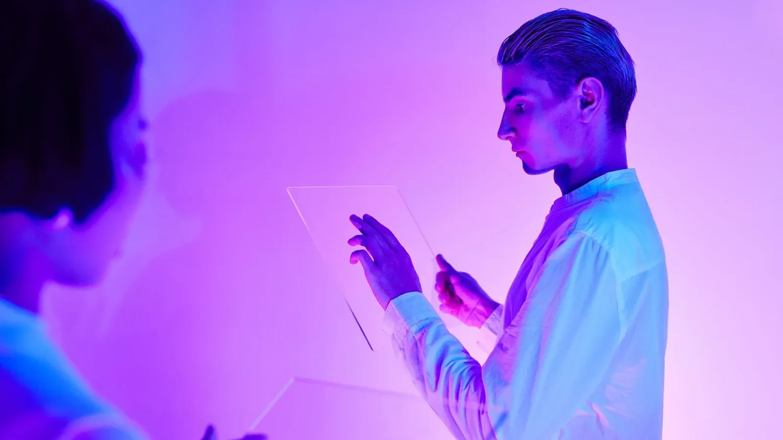 Two individuals engaging with futuristic transparent touch screens in a vibrant purple-lit studio.