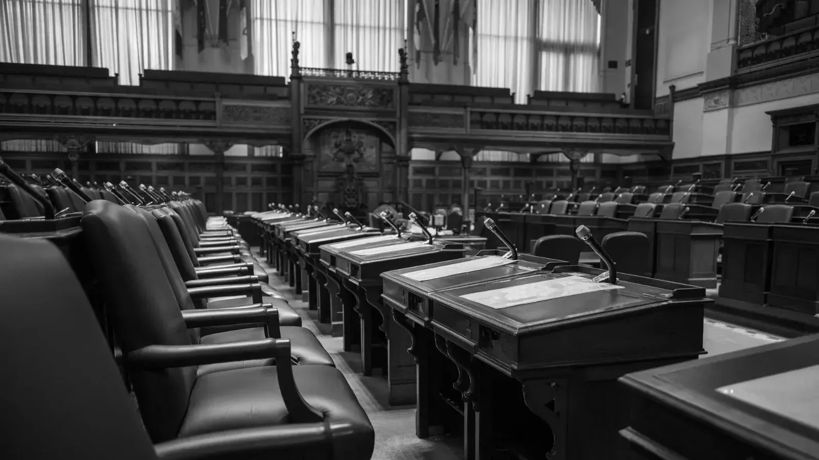 Foto en blanco y negro de una cámara de asamblea legislativa tradicional, con asientos y escritorios vacíos.