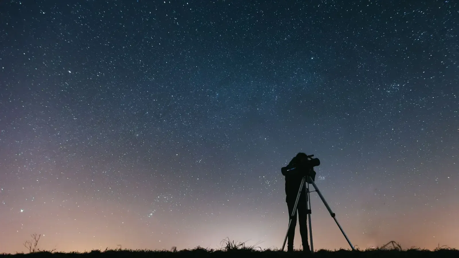 A silhouetted person using a telescope to observe the night sky filled with stars.