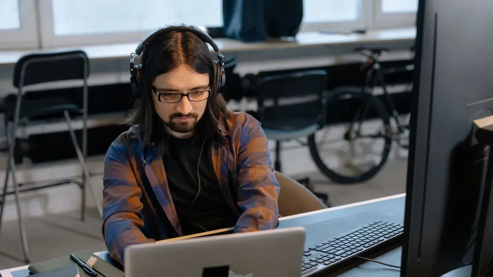 Desarrollador masculino barbudo en auriculares que codifican en una computadora portátil en un entorno de oficina moderno.