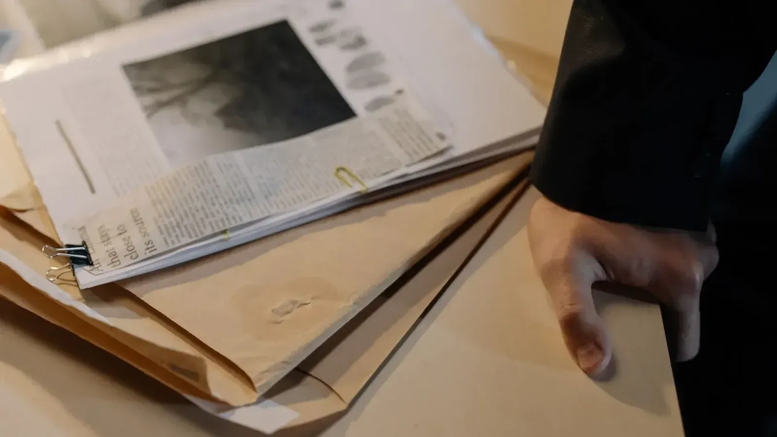 Close-up de una mano descansando sobre un escritorio cerca de una pila de documentos de investigación y recortes de periódicos.