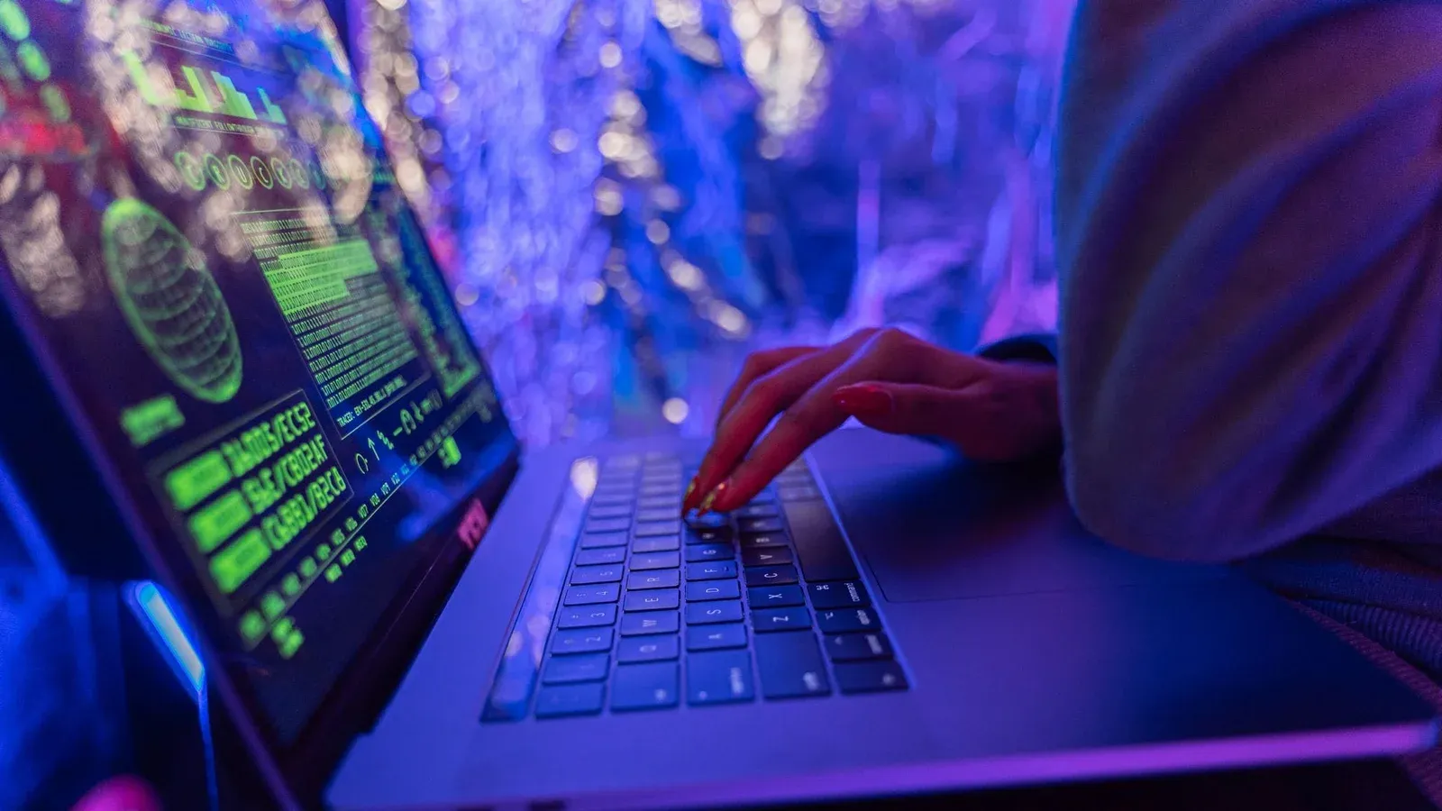 Close-up of hands typing on a laptop displaying cybersecurity graphics, illuminated by purple light.