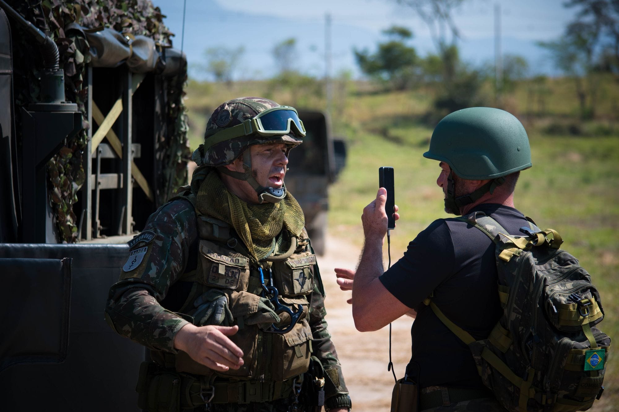 Two soldiers in full gear having a tactical discussion outdoors beside military vehicles.