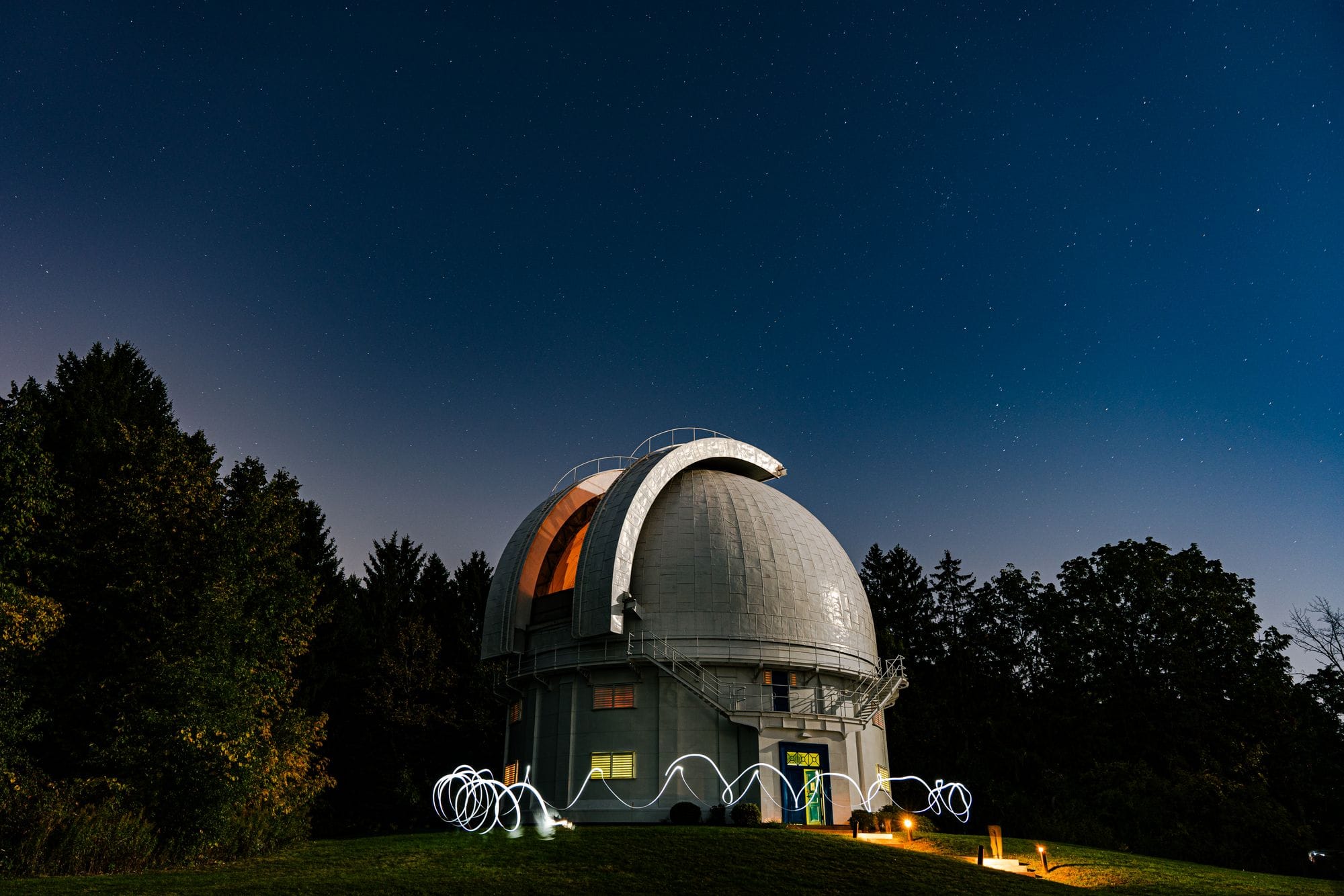 A scenic view of an observatory with a star-filled sky at night, surrounded by trees.