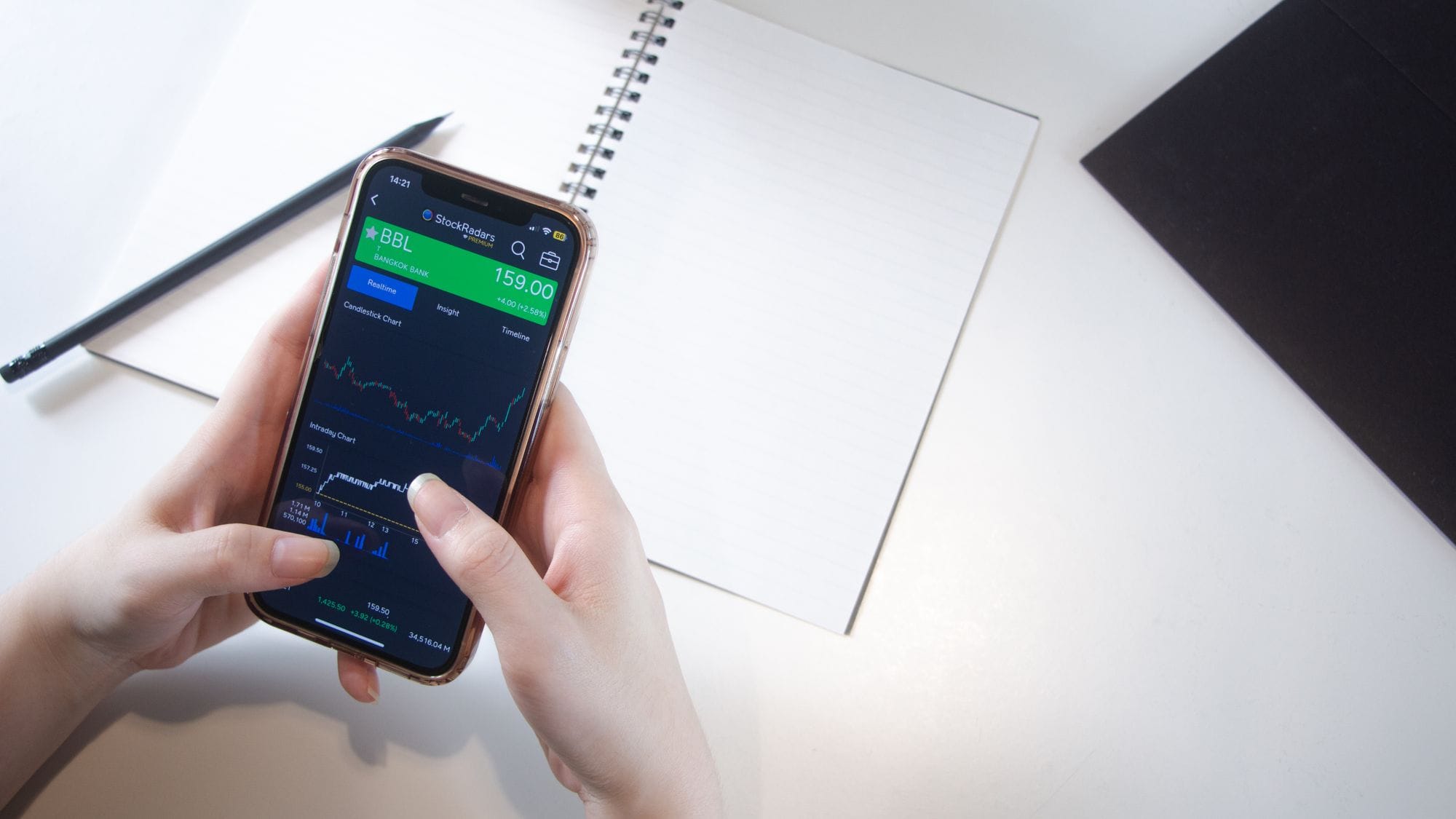 Close-up of hands using smartphone stock market app with notebook and pencil on desk.