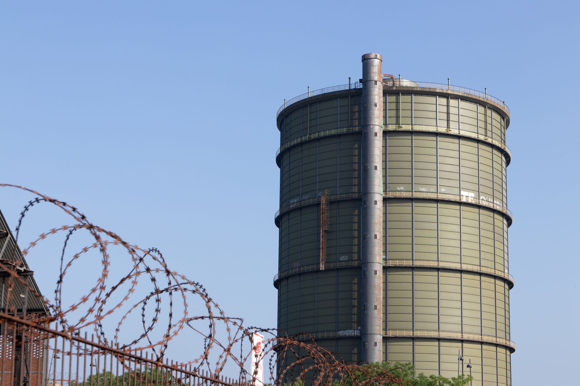 A large gas holder in an industrial area surrounded by barbed wire under a clear blue sky.