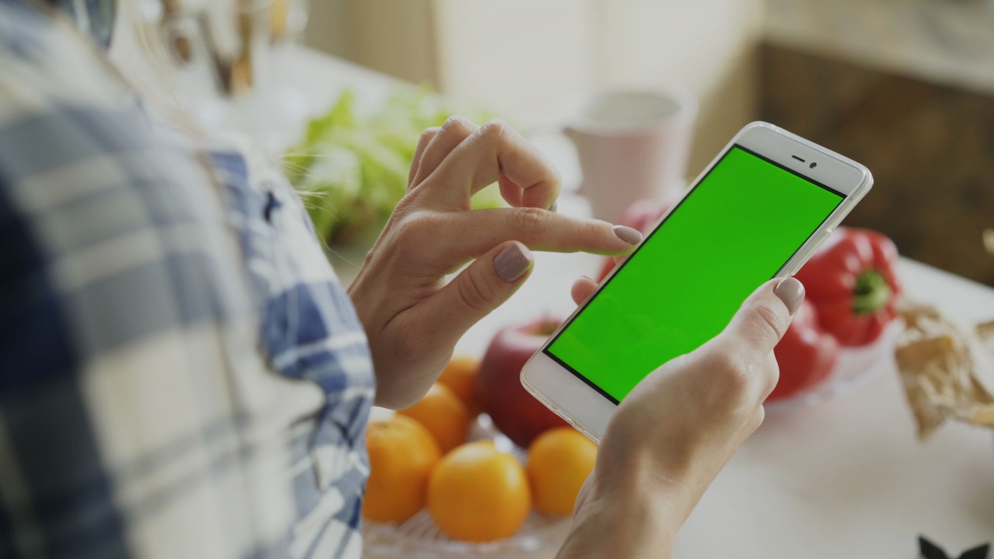 A person using a smartphone with a green screen surrounded by fresh fruits and vegetables in the kitchen.
