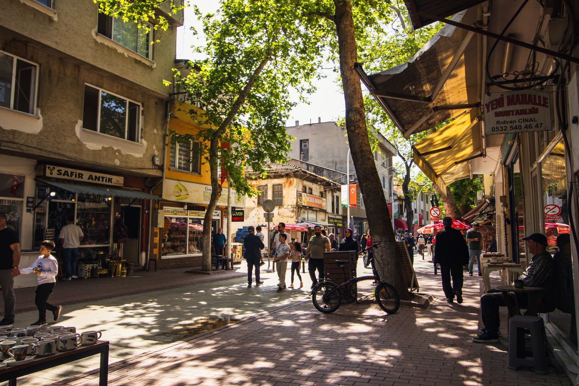 Busy street with people, shops and trees in a lively urban neighborhood during the day.