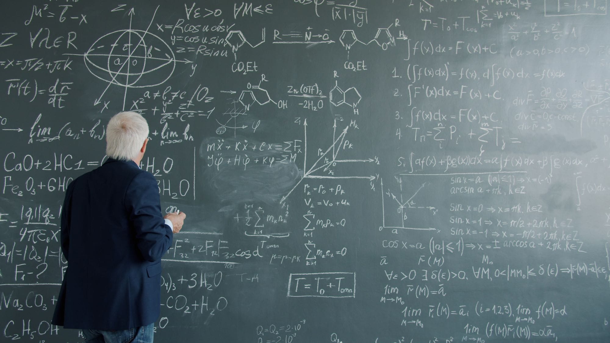 A mature professor writes complex mathematical equations on a large chalkboard.