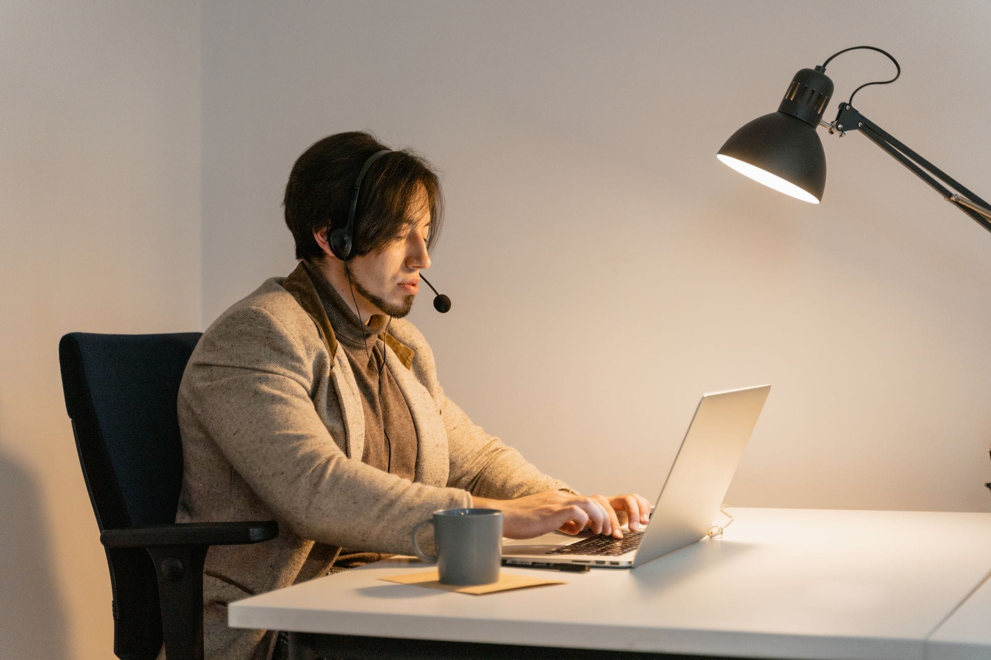 Professional man in a cozy office setting using a laptop for work-related tasks, wearing a headset under a desk lamp.