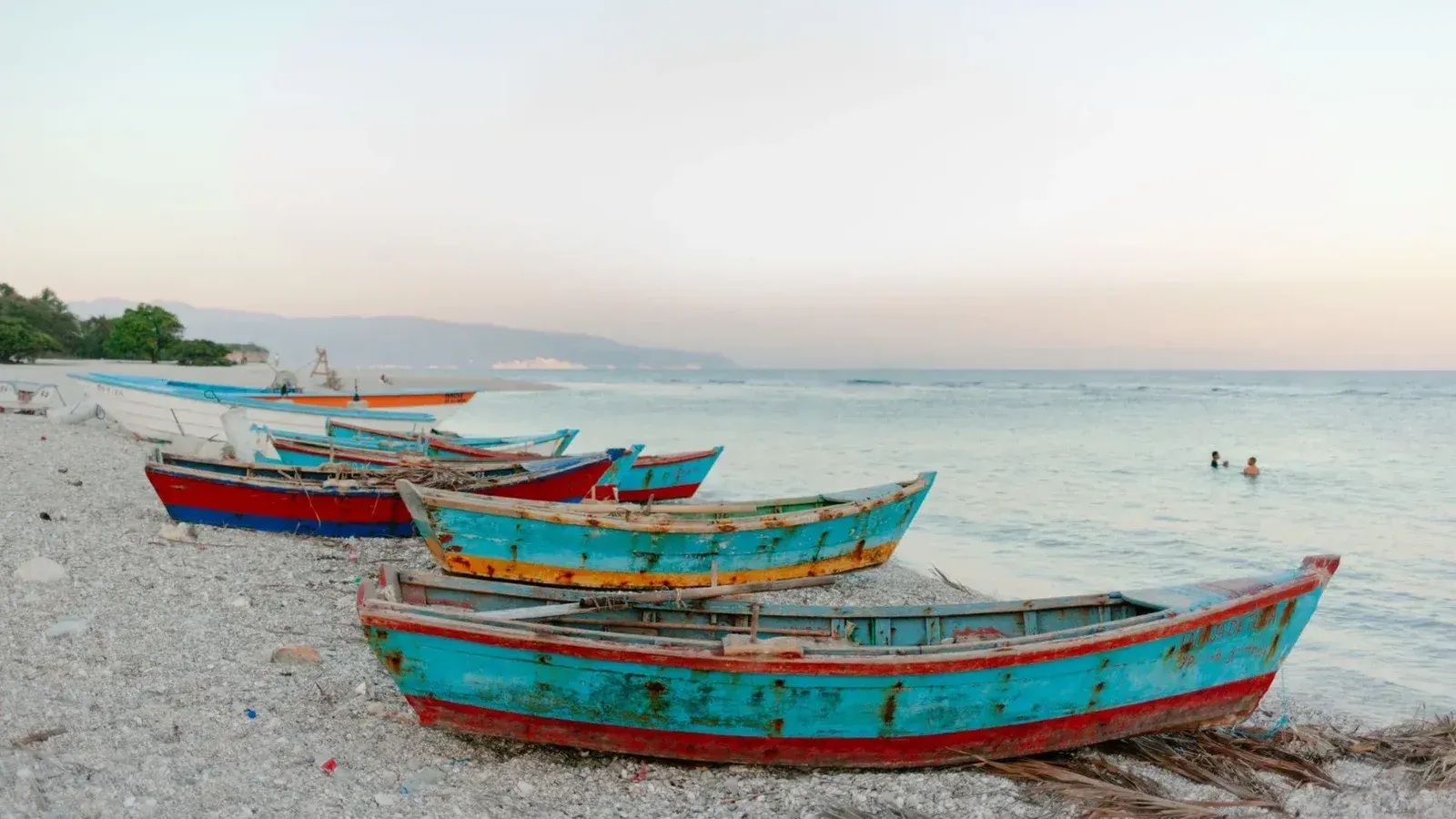 Shabby wooden fishing boats moored on pebble coast near travelers swimming in rippling sea in evening