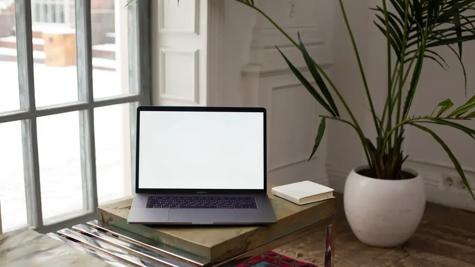 A minimalist indoor scene features a laptop, book, and potted plant on a wooden table by a window.