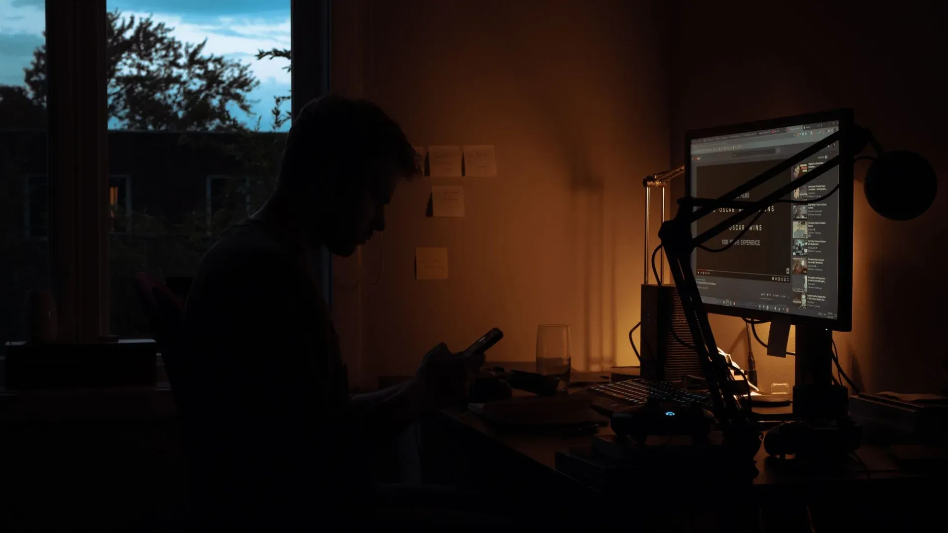 Silhouette of a man using a smartphone in a dark room with computer screen lighting.