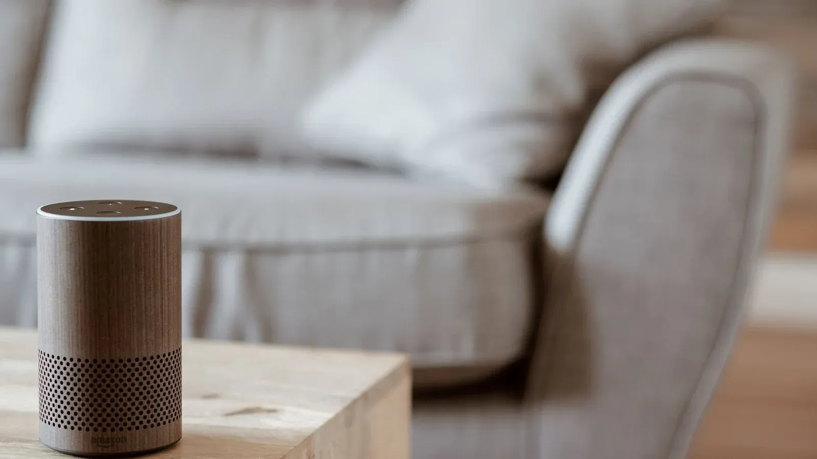 A modern smart speaker sits on a wooden table in a cozy, minimalist living room setting.