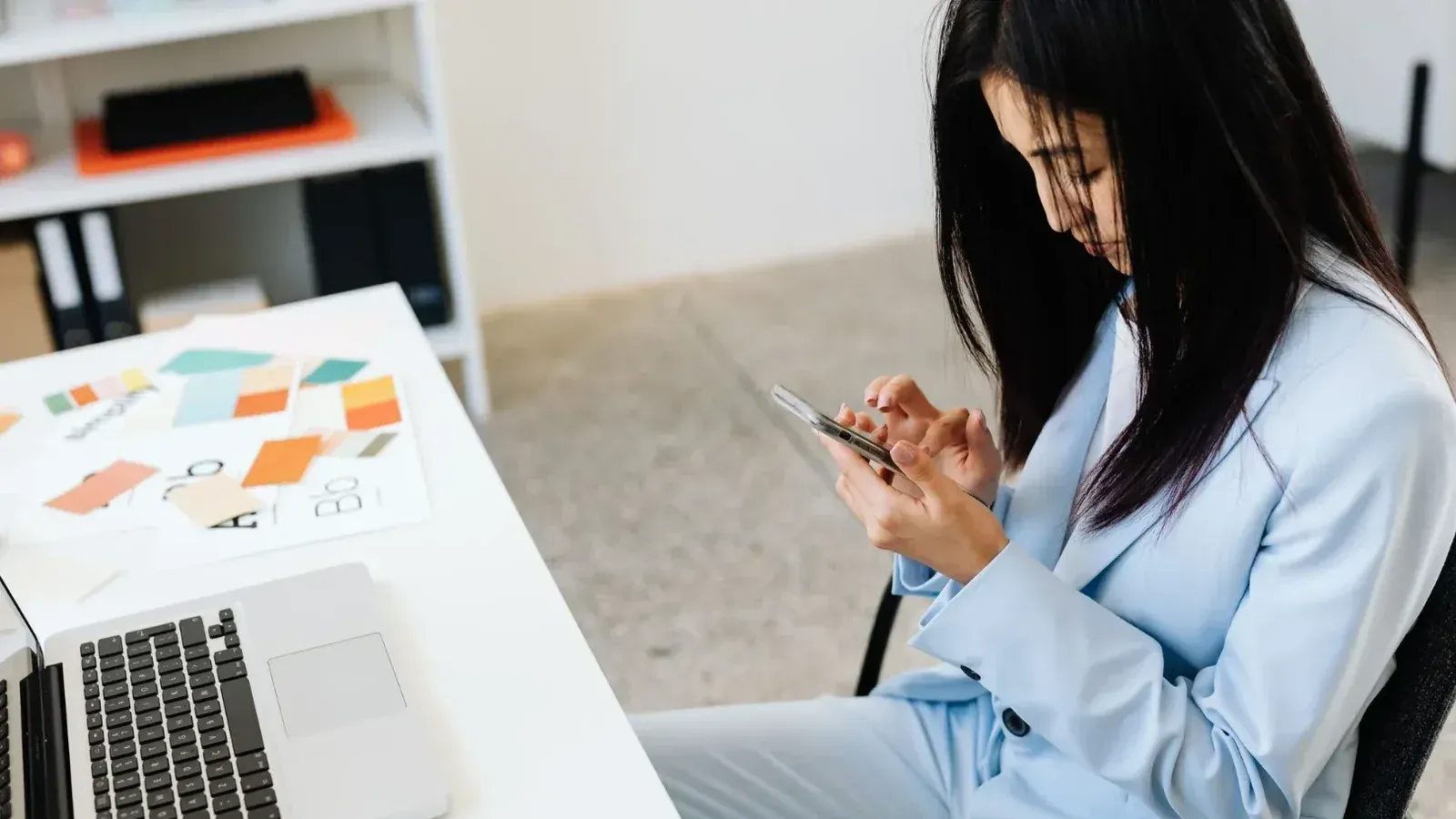 Mujer asiática en un traje azul con smartphone en el escritorio de oficina con muestras de ordenador portátil y de color.