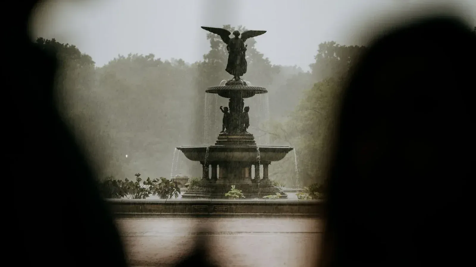 A beautiful angel statue fountain in a lush park during rainfall, creating a serene ambiance.