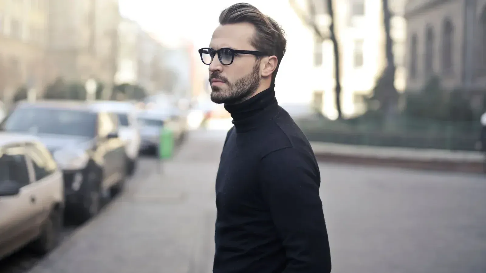 A fashionable man in a black top and glasses poses in an urban street with parked cars.