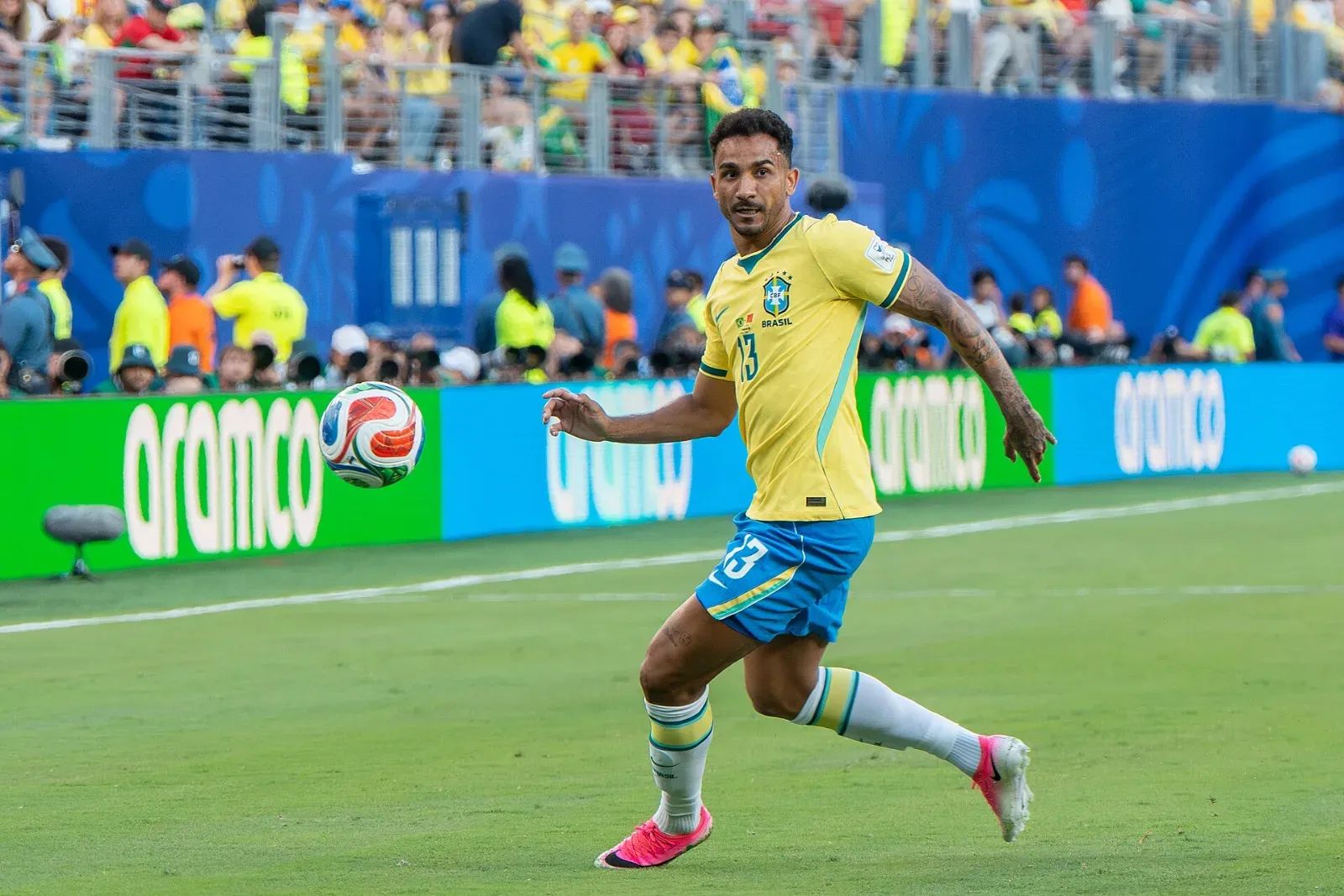 Danilo, con la camiseta amarilla número 13 de Brasil y short azul, corre hacia el balón cerca de la banda, con fotógrafos y aficionados al fondo.