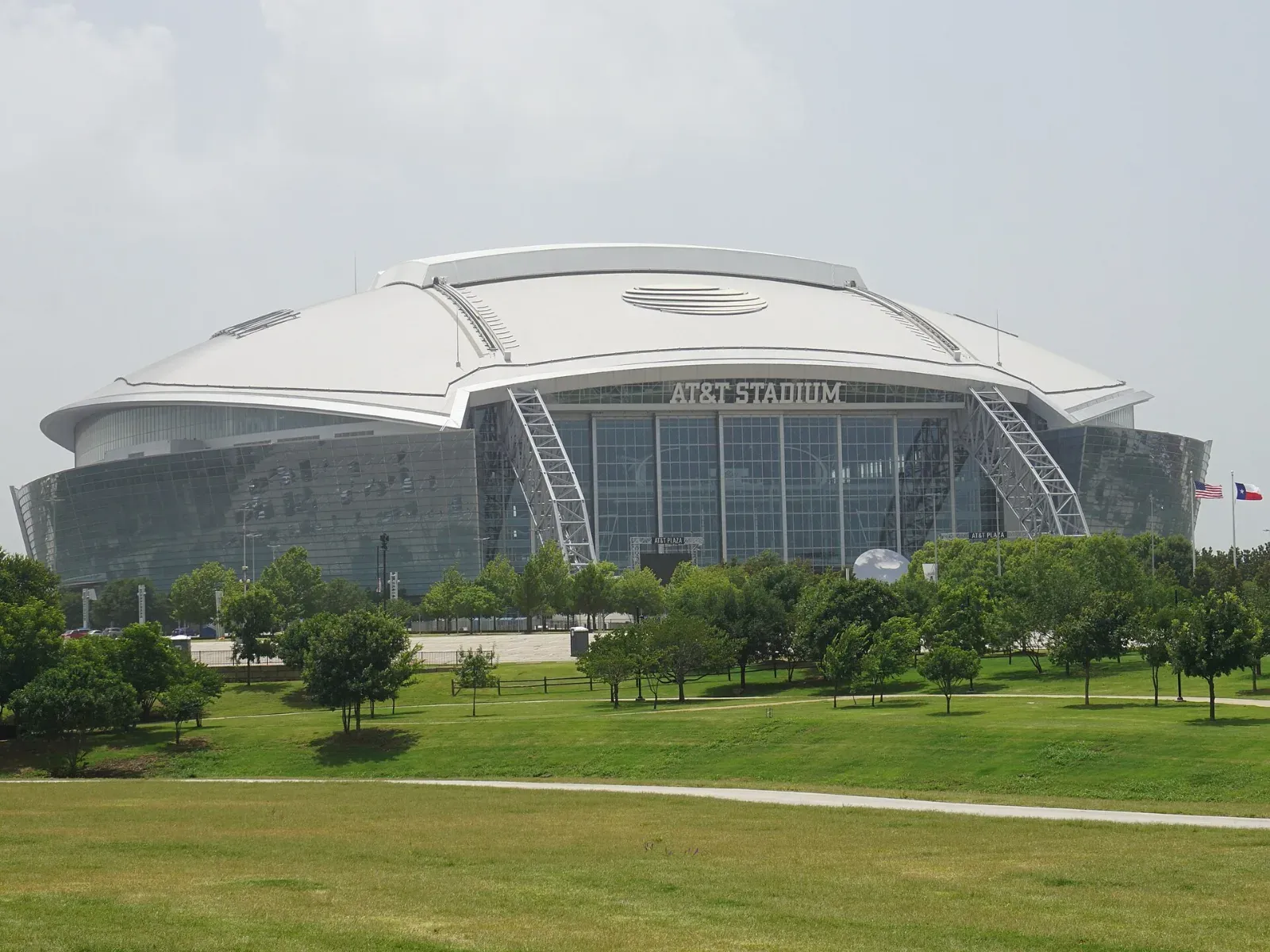 Exterior del AT&T Stadium, un estadio de techo abombado y fachada de vidrio con su nombre en lo alto, rodeado de césped y árboles bajo un cielo nublado.