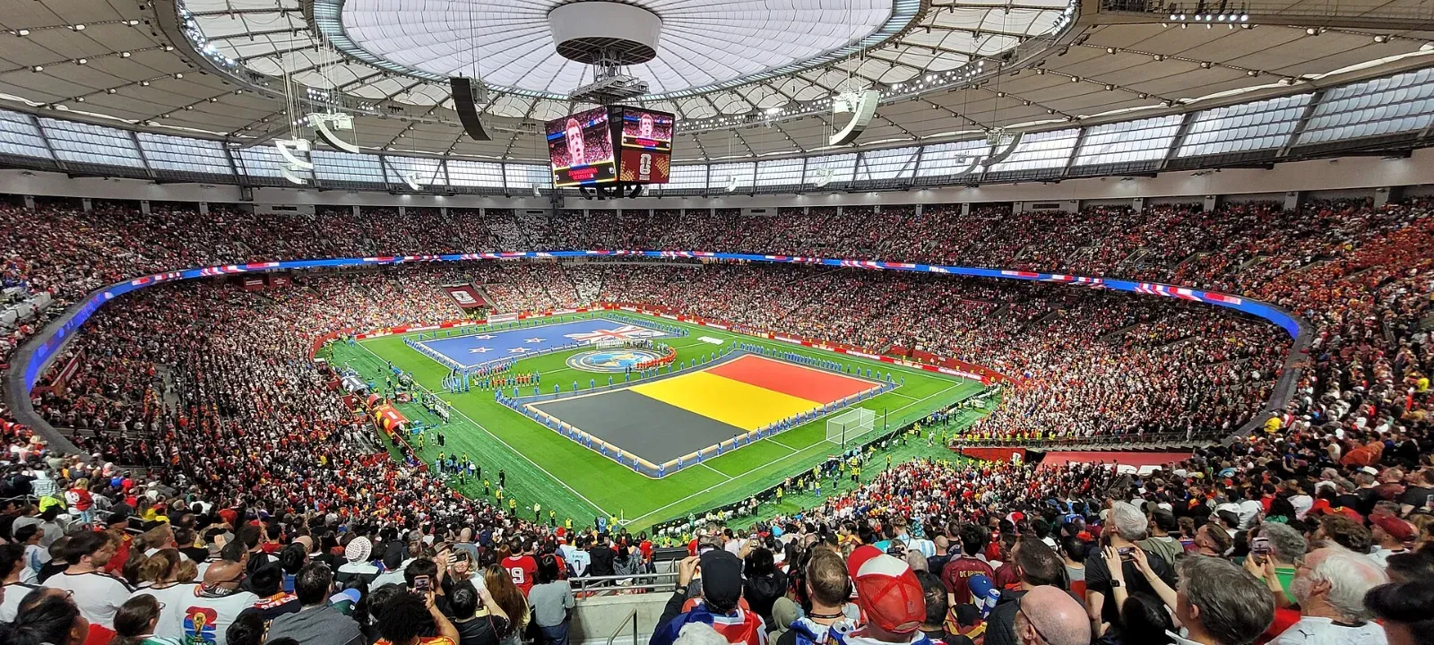 Estadio techado y lleno, con dos banderas gigantes extendidas sobre el césped, la de Bélgica y la de Nueva Zelanda, durante la ceremonia previa a un partido.