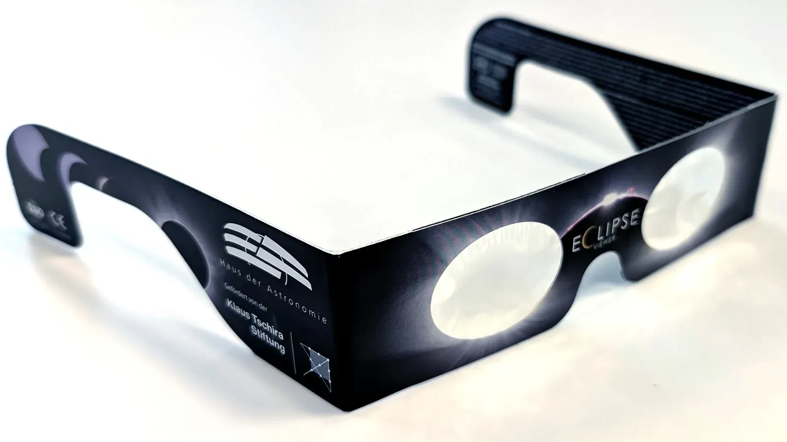 Gafas de cartón para observar eclipses, con filtros plateados, el logotipo de la Haus der Astronomie y la palabra «Eclipse» impresos, sobre una superficie blanca.