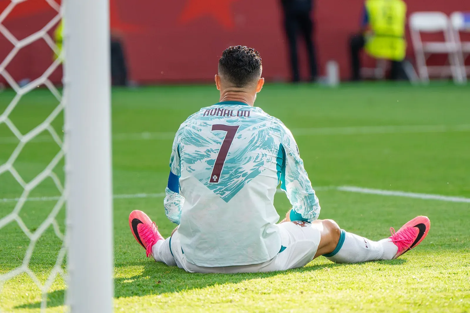 Cristiano Ronaldo, de espaldas con el dorsal 7 y la camiseta blanca y turquesa de Portugal, sentado en el césped junto al poste de una portería.