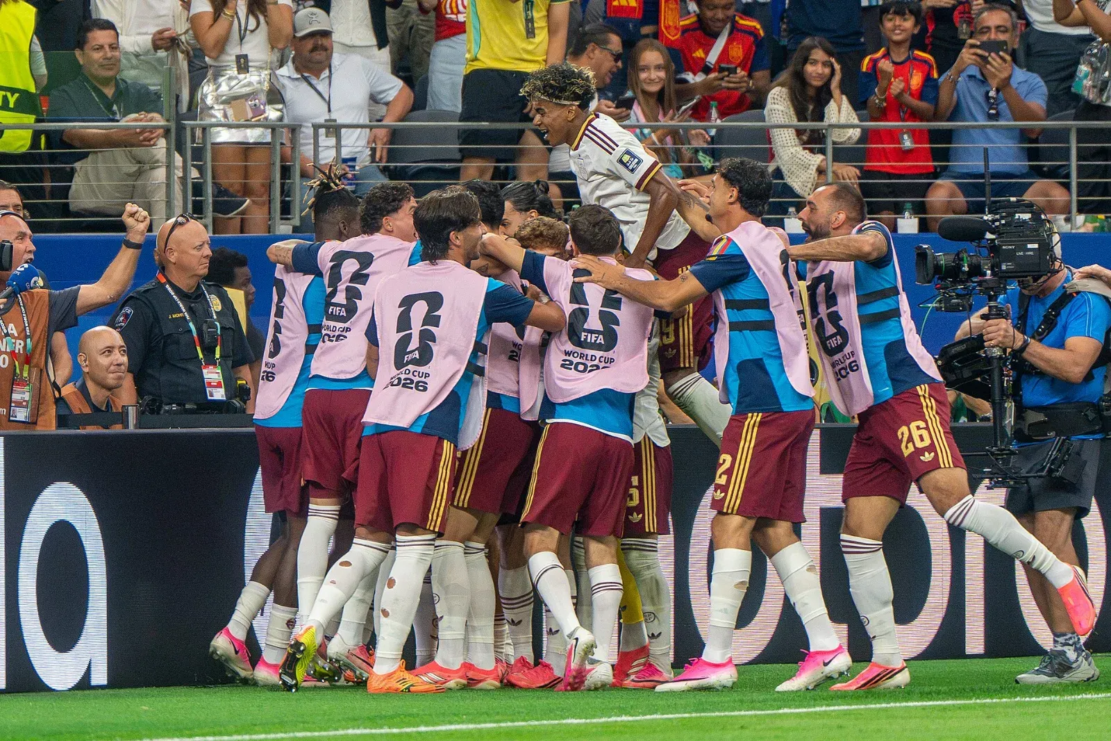 Jugadores de España celebran abrazados junto a la banda; varios llevan petos rosas del Mundial 2026 y uno de camiseta blanca salta sobre el grupo, con aficionados en la grada.