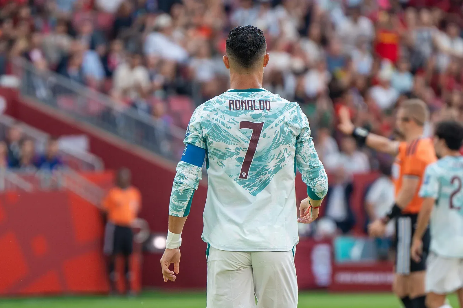 Cristiano Ronaldo, de espaldas, con la camiseta blanca y turquesa de Portugal con su nombre, el número 7 y el brazalete de capitán; al fondo, la grada desenfocada.