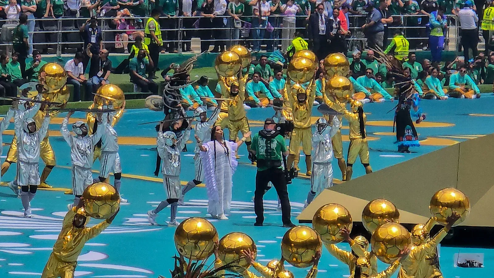 Lila Downs, con vestido blanco, en el centro del campo entre bailarines vestidos de blanco, danzantes con penachos de plumas y figuras doradas que sostienen balones dorados.