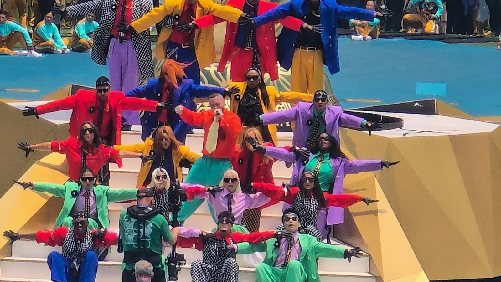 J Balvin, con chaqueta roja y pantalón turquesa, canta con un micrófono en una escalinata rodeado de bailarines con trajes de colores y gafas oscuras.