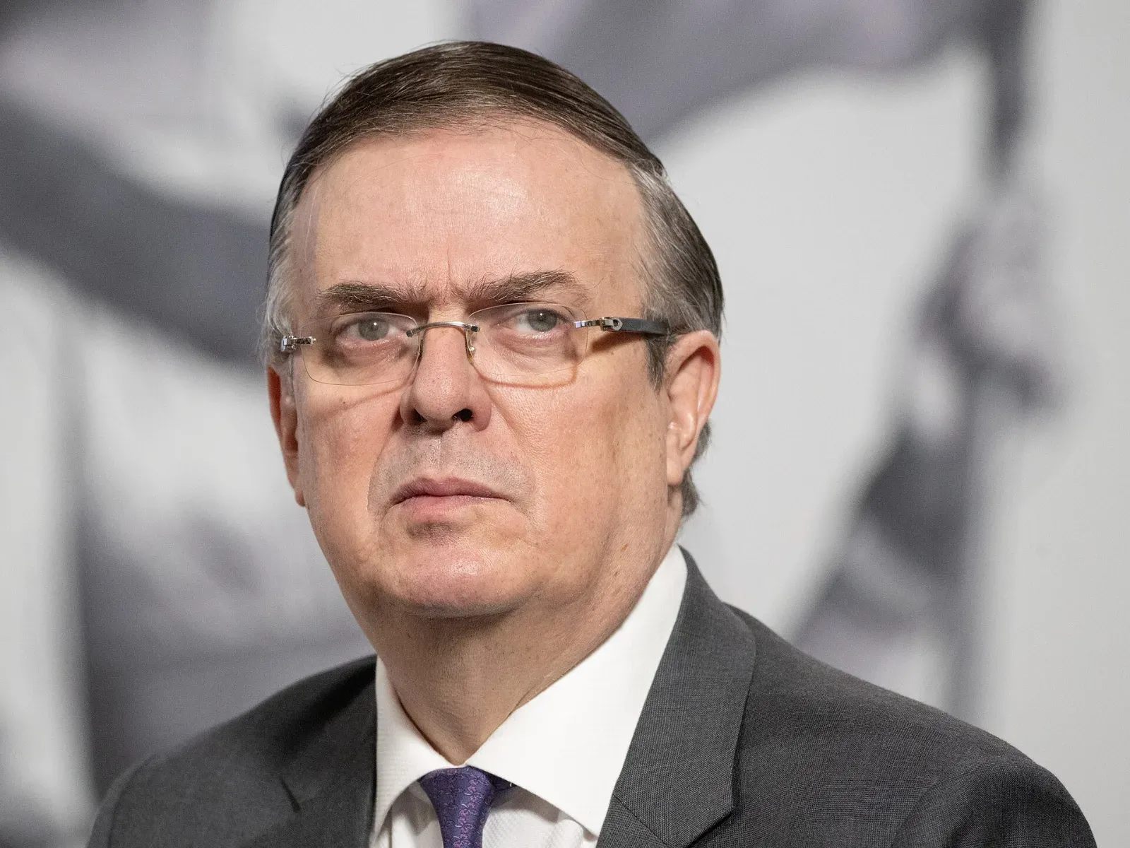 Marcelo Ebrard, con anteojos, traje gris oscuro, camisa blanca y corbata morada, mira hacia un lado frente a un fondo claro.