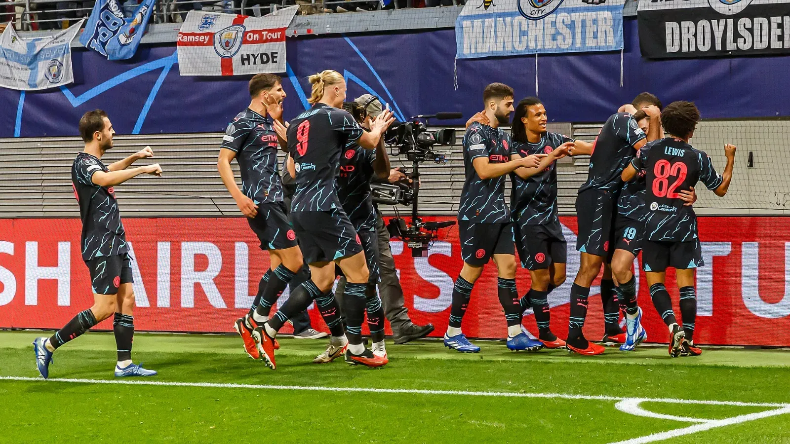 Jugadores del Manchester City, con uniforme azul oscuro, celebran un gol junto a la banda, frente a banderas de aficionados con la palabra «Manchester».