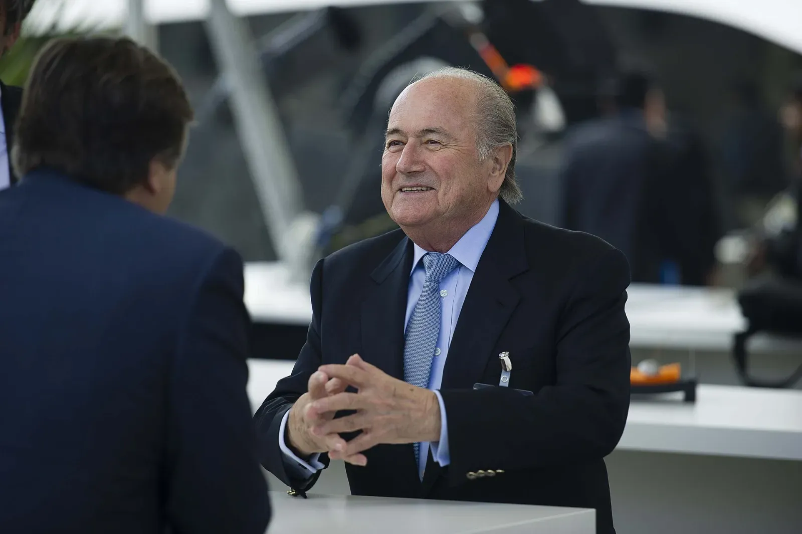 Joseph Blatter, de traje oscuro y corbata celeste, sonríe con las manos entrelazadas apoyado en una mesa alta mientras conversa con un hombre de espaldas.