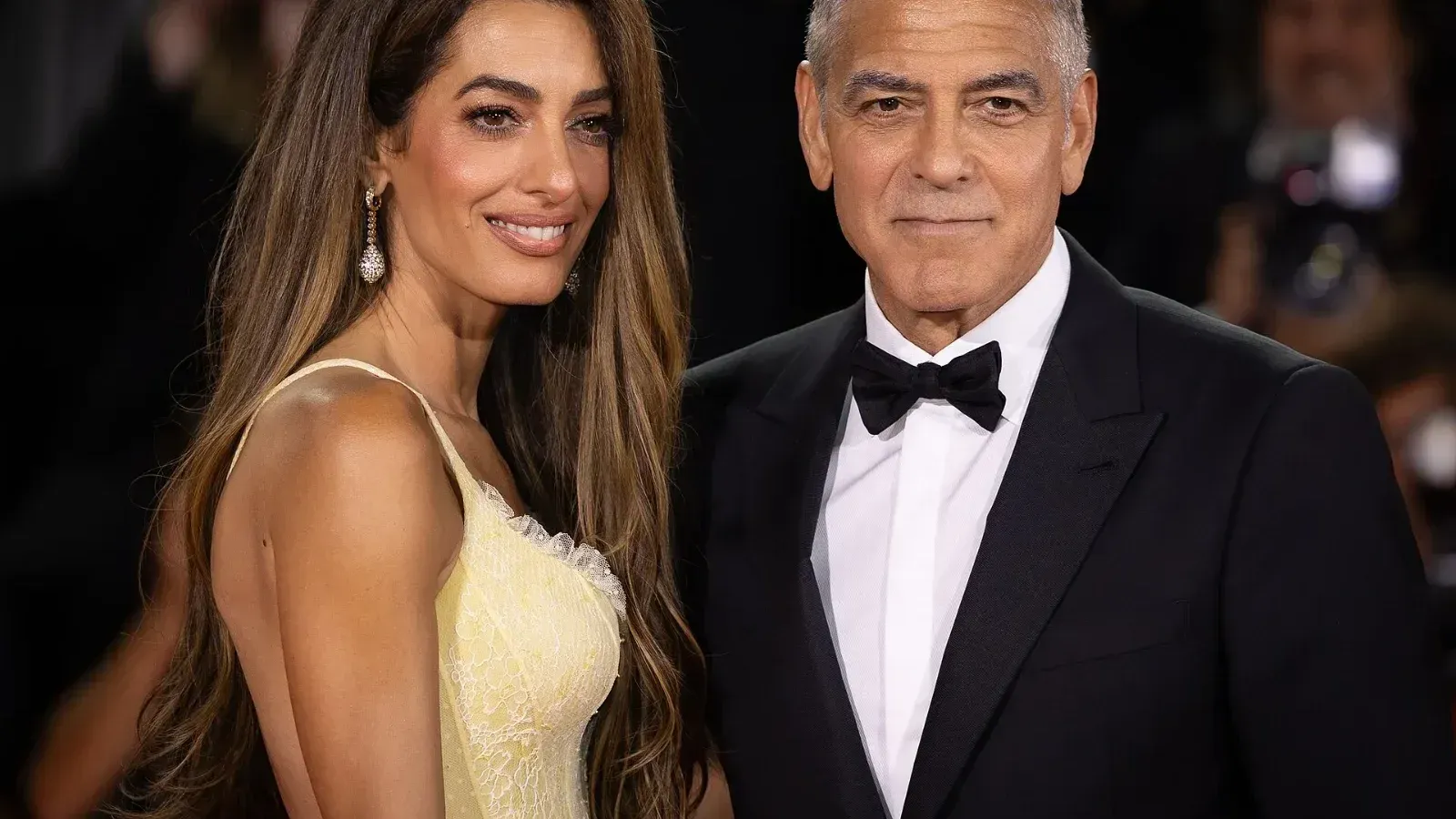 Amal Clooney, con un vestido amarillo claro de tirantes, posa junto a George Clooney, de esmoquin y pajarita negra, con fotógrafos al fondo en el Festival de Venecia.