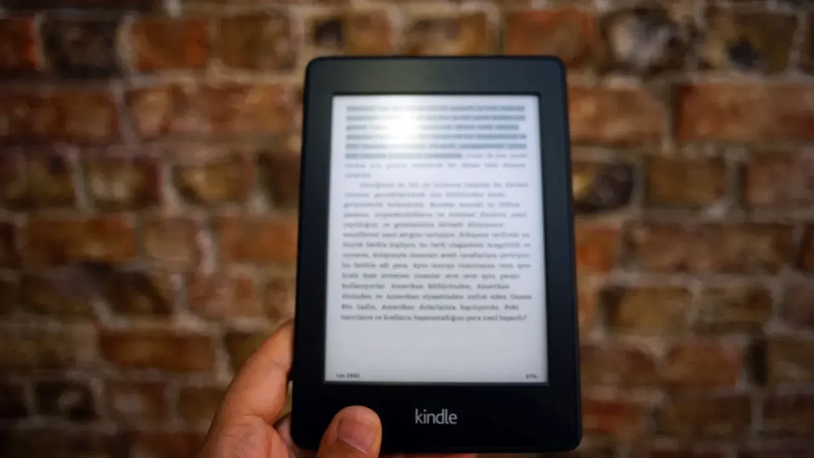 Close-up of a Kindle e-reader held by a person, showcasing a digital reading device against a textured brick wall backdrop.