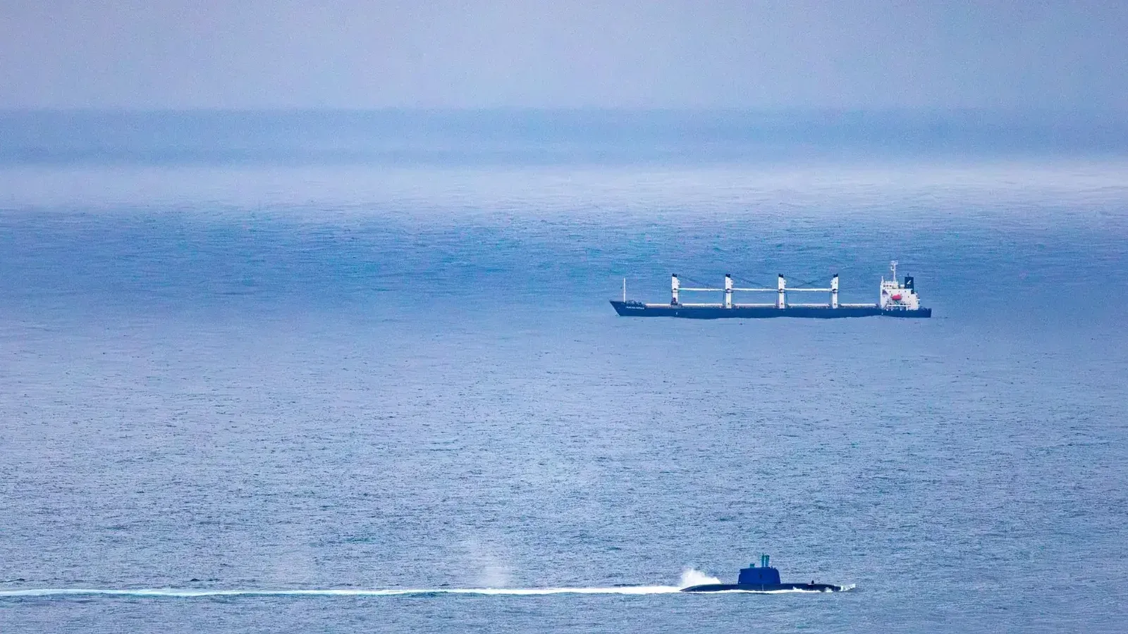Un barco de carga y un submarino navegan por el vasto océano abierto bajo un cielo despejado.