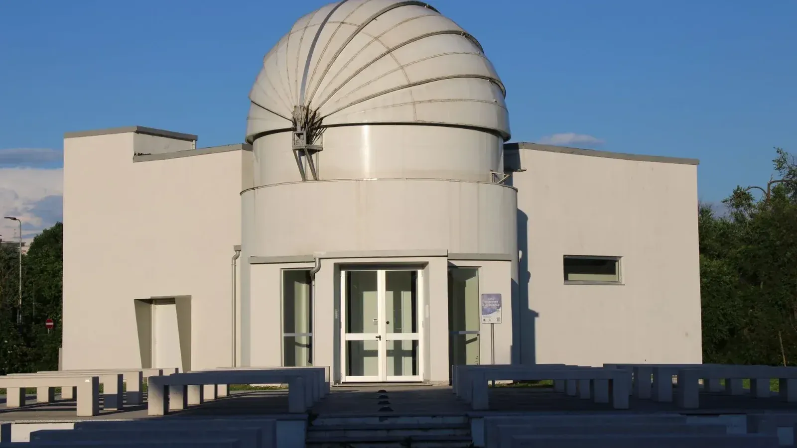 A white domed astronomical observatory in Milan surrounded by greenery under a clear blue sky.